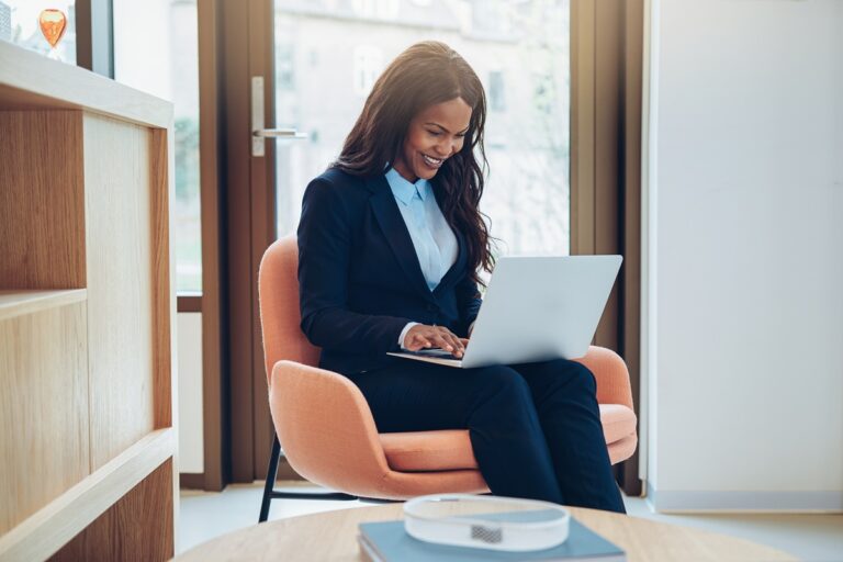 Smiling young African American businesswoman sitting in a chair in the lounge of a bright modern office working online with a laptop